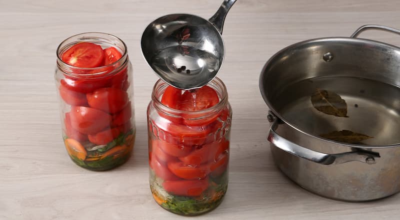 Разложите по стерилизованным банкам укроп и петрушку, колечки репчатого лука, кружки моркови и чеснок. Равномерно распределите растительное масло. Помидоры вымойте, разрежьте на половинки и удалите основания плодоножек.