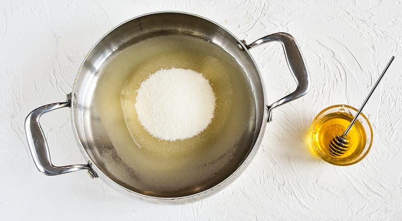 Налейте на дно толстодонной кастрюли 150 мл воды и мед. Насыпьте сахар и, не перемешивая, поставьте на средний огонь. Иногда немного покачивайте кастрюлю, чтобы сахар таял более равномерно. Когда весь сахар растает, поварите сироп 5–7 мин. и снимите с огн