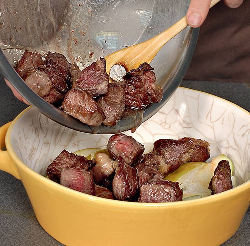 Говядину нарезать кубиками со стороной 3 см и обжарить в разогретом масле, 6 мин. Переложить в форму для запекания. В сотейник, где жарилось мясо, положить лук и готовить 4 мин.