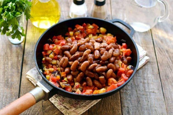 Фасоль после варки промываем, воду сливаем, фасоль добавляем к овощам. Держим на огне еще около 7 минут. Отправляем в сковороду грецкие орехи, чеснок, перец, хмели-сунели, зелень, соль. По вкусу можно добавить и иные специи. Перемешиваем массу на сковород