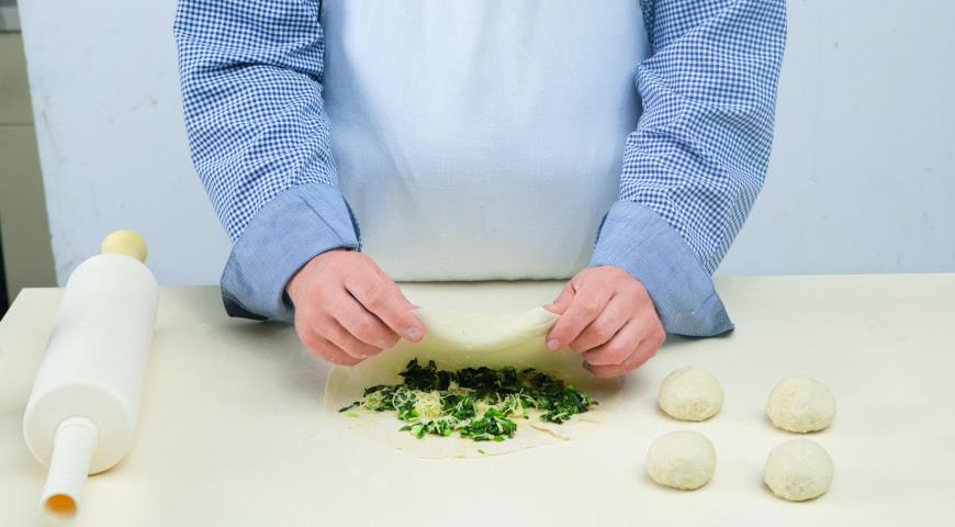 В остывшую начинку всыпьте отложенный сыр. Тесто разделите на равные части весом примерно по 40 г. Каждый кусочек раскатайте в лепешку, посыпьте зеленью, сложите пополам, защипните края и снова аккуратно раскатайте.
